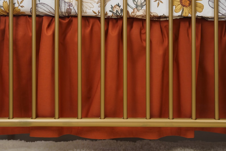 Op de afbeelding: Een roestkleurige wieg rok met een gerimpelde rand hangt aan een gouden metalen wieg frame.