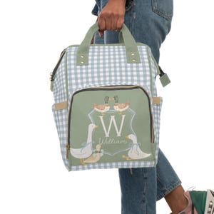 May include: A blue and white gingham diaper bag with a green patch featuring a monogram of the letter W and the name William. The patch also has a design of three ducks.