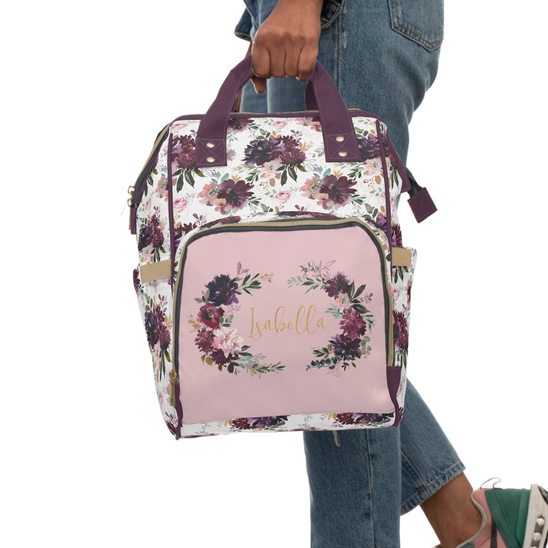May include: A pink and white floral print diaper bag backpack with a pink front pocket featuring a floral wreath and the name "Isabella" in gold lettering.