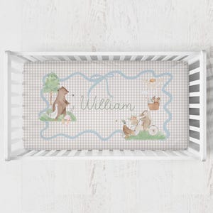 May include: A crib sheet with a light gray and white gingham pattern. The sheet features a blue ribbon border with the name "William" in cursive. Illustrations include a bear, goose, rabbit, and a mouse in a basket.