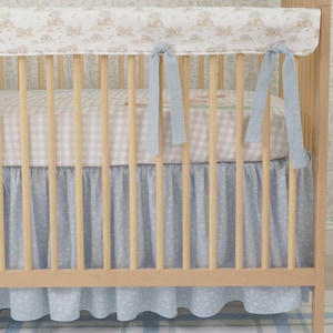Op de afbeelding: Een houten babybedje met een lichtblauwe en witte bloemenpatroon bedrok en bumper. Het beddengoed omvat een geruit laken en lichtblauwe linten. Het bedje is gemaakt van lichtgekleurd hout.