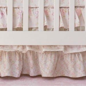Könnte beinhalten: Ein weißer Gitterbettrahmen verdeckt teilweise einen gerüschten Bettrüschenstoff. Der Stoff zeigt ein hellrosa Blumenmuster mit kleinen Rosen, grünen Blättern und zarten rosa Schleifen auf hellem Grund, was für eine geschichtete Optik sorgt.