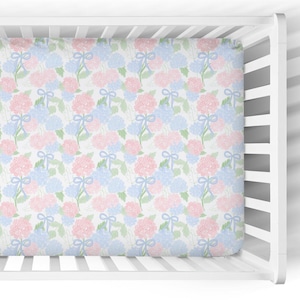 May include: A fitted crib sheet with a floral pattern of pink and blue hydrangeas and light blue bows on a white background. The sheet is made of soft fabric and is designed to fit a standard crib mattress.