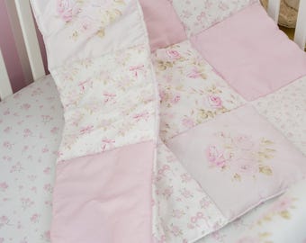 May include: A quilted baby blanket featuring a patchwork design of solid light pink fabric and white fabric printed with pink roses and small bows. The blanket is displayed on a matching white sheet with a delicate pink floral pattern.