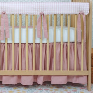 May include: A light wood crib with a pink gingham bumper and a matching pink crib skirt. The bumper is tied to the crib with pink ribbons. The crib skirt has a pleated design.
