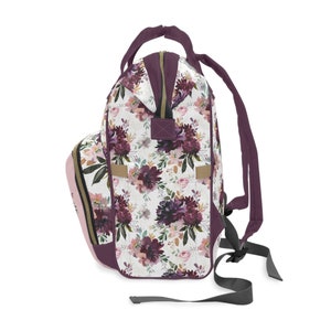 May include: A purple and pink floral print backpack with a zipper closure and adjustable straps. The backpack has a white background with a floral pattern in shades of purple, pink, and gold.