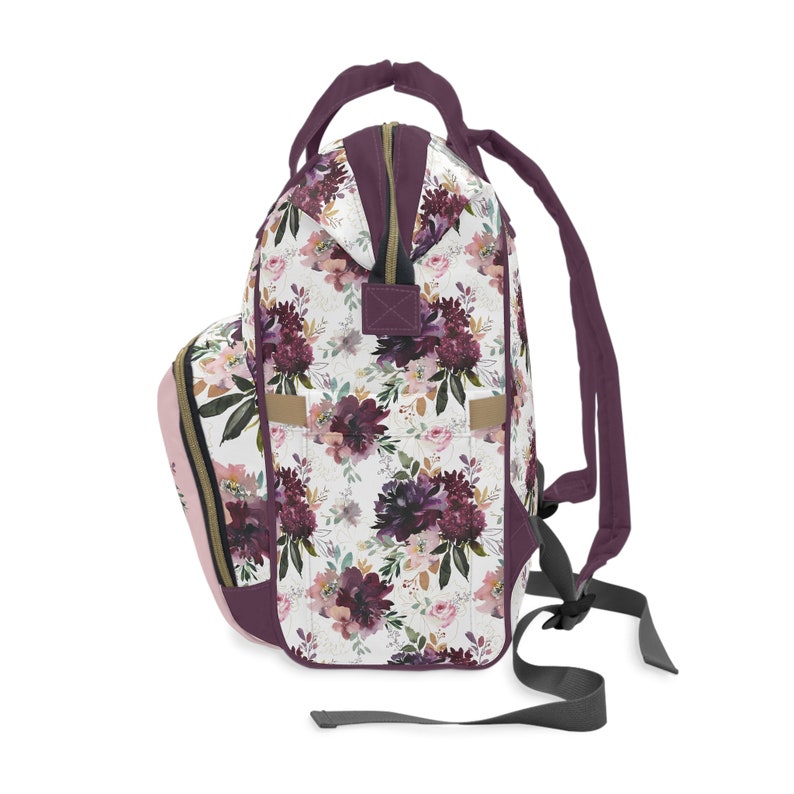 May include: A purple and pink floral print backpack with a zipper closure and adjustable straps. The backpack has a white background with a floral pattern in shades of purple, pink, and gold.
