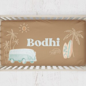 May include: A tan crib sheet with a light blue and white illustration of a vintage van, palm trees, a surfboard, and the name "Bodhi".