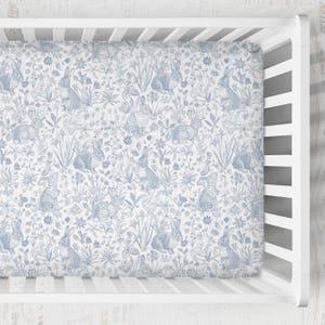 May include: A fitted crib sheet with a blue and white floral and bunny rabbit pattern. The sheet is made of soft fabric and is designed to fit a standard crib mattress. The overall theme is a whimsical nursery design.