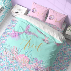 May include: A turquoise blue mermaid bedding set with a pink mermaid tail, seashells, and starfish. The set includes a duvet cover, two pillowcases, and a decorative throw pillow. The pillowcases and throw pillow feature the name "Ariel" in pink script.