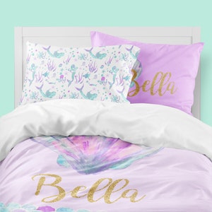 May include: A purple and white bedding set with a mermaid and seashell design. The bedding set includes a duvet cover, two pillowcases, and a decorative pillow. The decorative pillow has the name "Bella" written in gold glitter.