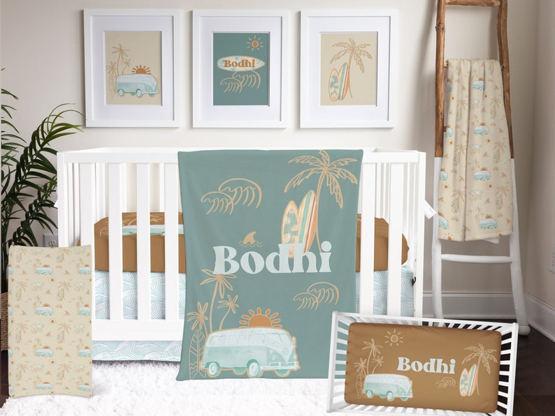 May include: A baby's nursery decorated in a beach theme. The crib is white with a brown and blue bedding set. There are three framed pictures on the wall, each with a different beach scene. The pictures are all in white frames. There is a brown and blue blanket on the crib, and a brown and blue pillow on the floor. The blanket and pillow have a beach scene with a van, palm trees, and surfboards. The name "Bodhi" is written on the blanket and pillow.