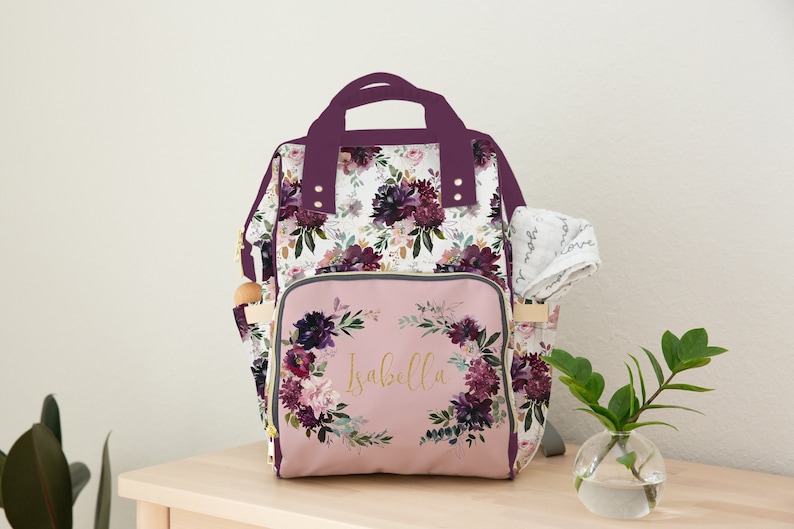 May include: A floral patterned diaper bag with a pink front pocket featuring a floral wreath and the name "Isabella" in gold lettering. The bag has a purple trim and gold accents.