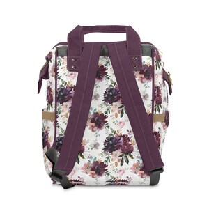 May include: Purple floral print backpack with adjustable straps and a top handle. The backpack has a white background with a floral pattern in shades of purple, pink, and green.