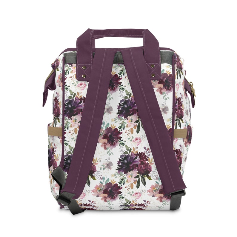 May include: Purple floral print backpack with adjustable straps and a top handle. The backpack has a white background with a floral pattern in shades of purple, pink, and green.