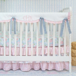 May include: A white crib with a floral patterned sheet and pink ruffled skirt. The crib bumper is white with pink trim and blue ribbons. The initials "CHR" are embroidered on the bumper. The crib is set against a blue and white striped wallpaper.