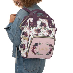 May include: A pink and purple floral diaper bag backpack with a gold zipper and a gold wreath with the name "Isabella" on the front.