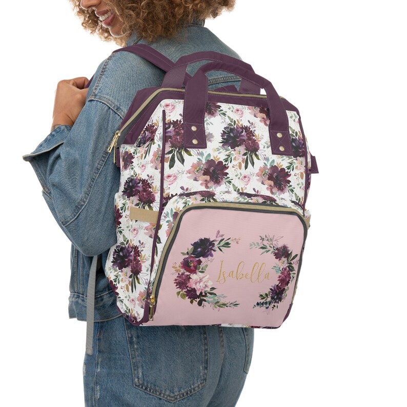 May include: A pink and purple floral diaper bag backpack with a gold zipper and a gold wreath with the name "Isabella" on the front.