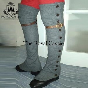 May include: Grey over-the-knee boot covers featuring a vertical row of dark buttons along the outer side and a brown leather strap with a metal buckle across the mid-calf. The covers are worn over black shoes and red trousers, displaying a textured fabric.