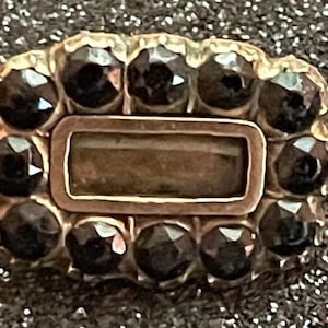 May include: A vintage gold-toned brooch with a rectangular center and a border of dark, faceted gemstones. The brooch has a rectangular center and a pin on the back. The gemstones are dark and reflect light.