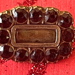 May include: A vintage rectangular brooch with a gold-toned frame. The brooch features a central rectangular element and is surrounded by dark, round gemstones. The background is a textured red.