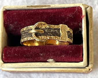 Puede incluir: Un anillo dorado con forma de hebilla de cinturón, con una banda gris oscuro. El anillo se presenta en una pequeña caja rectangular con interior de terciopelo rojo.