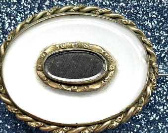 May include: A white oval cameo brooch with a gold-toned twisted rope border and a smaller oval mirror inset in the center.