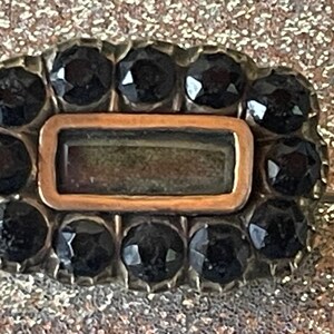 May include: Rectangular antique brooch with a central copper-colored rectangle and a border of black gemstones. The brooch has a silver-colored metal setting and a pin on the back. The background is a glittery gold color.