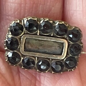 May include: Antique rectangular brooch with a gold-toned frame and a central rectangular stone. The brooch is surrounded by a border of dark, faceted stones. The pin is set against a skin-toned background.
