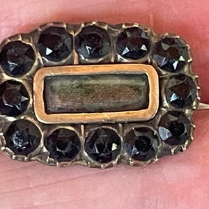 May include: Antique rectangular brooch with a gold-tone frame and a central rectangular element. The brooch is surrounded by a border of dark, faceted stones. A pin is attached to the back.