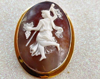May include: A cameo brooch featuring a white carved figure of a woman holding a staff and grapes, set in a gold tone oval frame.