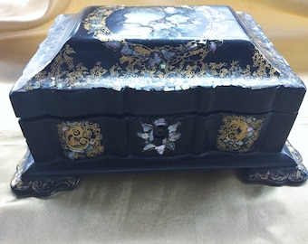 May include: A black wooden box with gold and iridescent accents. The box has a hinged lid and sits on four feet. The box is decorated with floral designs and intricate patterns.