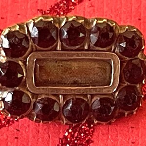 May include: A rectangular antique brooch with a gold-tone frame. The brooch is adorned with dark, faceted gemstones around the perimeter and a central rectangular element. The gemstones are set closely together, creating a striking contrast against the gold setting.