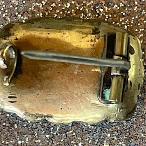 May include: Back view of a vintage gold-toned brooch with a pin closure. The brooch has a rectangular shape with a slightly uneven edge. The pin is silver-colored and attached to the back with a clasp.