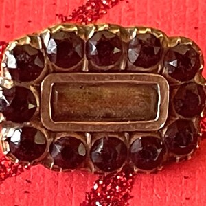 May include: A vintage rectangular brooch with a gold-tone frame. The brooch is adorned with dark, round gemstones surrounding a central rectangular stone. The gemstones are set closely together, creating a sparkling effect. The brooch is set against a red background.