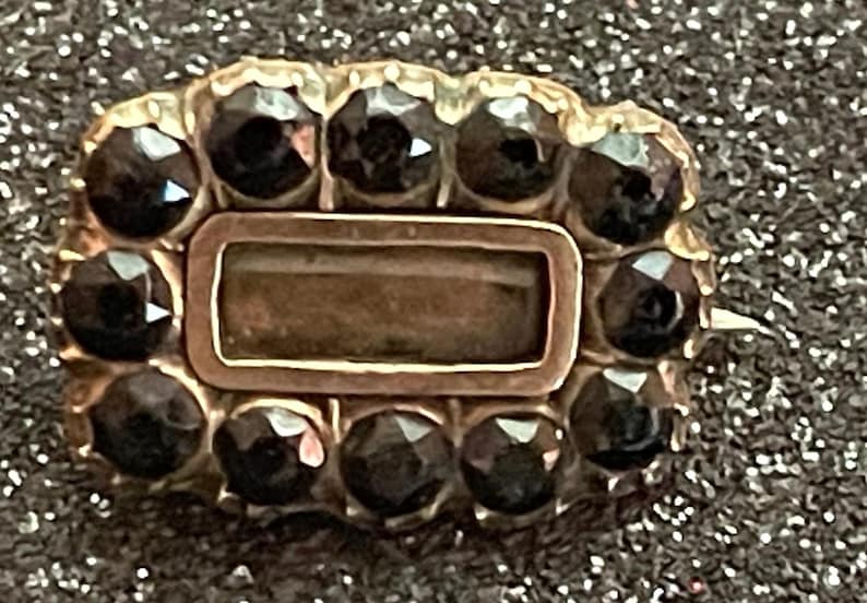 May include: A vintage gold-toned brooch with a rectangular center and a border of dark, faceted gemstones. The brooch has a rectangular center and a pin on the back. The gemstones are dark and reflect light.