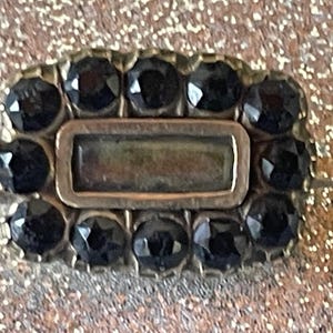May include: A rectangular, antique-style brooch with a silver-toned frame. The brooch is adorned with a border of black, faceted gemstones surrounding a central, rectangular, clear stone.