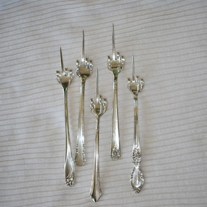 May include: Five silver forks with a middle finger design. The forks are arranged in a row on a white textured surface.