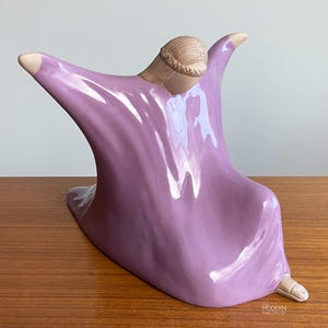 May include: A ceramic sculpture of a figure in a flowing lavender robe, arms outstretched. The figure's head and feet are a contrasting beige color. The sculpture is on a wooden surface.