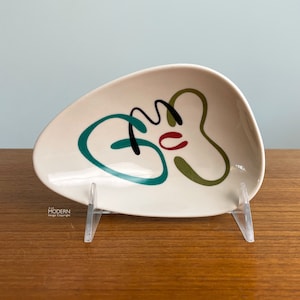 May include: Cream-colored, mid-century modern ceramic dish with abstract teal, black, red, and olive green design. The dish is teardrop-shaped and sits on a clear acrylic stand. A vintage decorative piece.