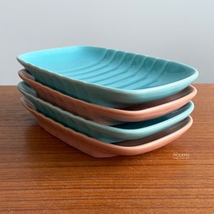 May include: A stack of four ceramic dishes in shades of turquoise and peach. Each dish has a rectangular shape with a curved edge and a ridged interior. The dishes are stacked on a wooden surface.