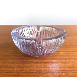 May include: A clear glass ashtray featuring a textured, ribbed exterior with vertical patterns and three distinct notches along the rim. The glass has a subtle iridescent sheen, reflecting light across its curved, bowl-shaped structure.