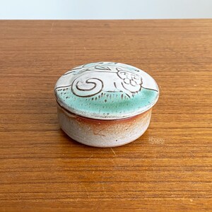 Puede incluir: Pequeña caja redonda de cerámica con tapa. La tapa está decorada con un diseño de gato blanco sobre un fondo turquesa. La caja tiene un cuerpo moteado en marrón claro y blanco. Sobre una superficie de madera.