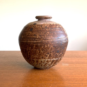 May include: A handcrafted, round ceramic vase with a textured, brown surface and a small, cylindrical neck. The vase features horizontal, etched lines and dark speckles, giving it a rustic appearance. The vase is displayed on a wooden surface.