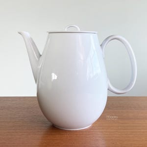 May include: A solid white porcelain teapot with a smooth, glossy finish. The teapot features a rounded, bulbous body, a curved spout, a C-shaped handle, and a flat lid with a small, arched handle on top. The design is clean and uniform.