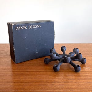 May include: A black, mid-century modern candle holder with a unique, spiky design. The candle holder is next to a dark blue box with the words "DANSK DESIGNS" printed on it. The items are on a wooden surface.