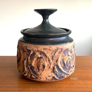 May include: A ceramic jar with a dark green lid and a textured, brown body. The body features a raised floral design. The jar sits on a wooden surface. The jar is a decorative storage container.
