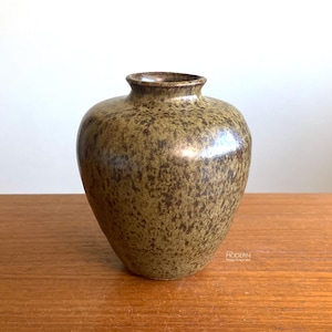 May include: A speckled, olive-green ceramic vase with a rounded body and a narrow neck. The vase has a textured surface with dark brown spots, and sits on a wooden surface. The vase is a decorative item.