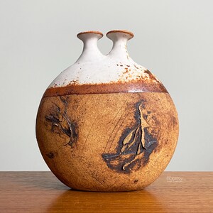 May include: A round ceramic vase with two narrow necks. The upper portion is finished in a smooth white glaze, while the lower body features a textured, earthy brown surface with dark, irregular markings and subtle scratch patterns throughout.
