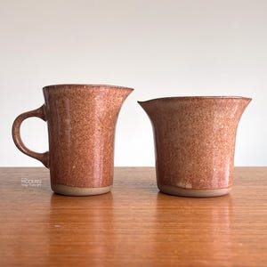 May include: Two speckled brown ceramic pitchers. One pitcher has a handle, and the other has a flared rim. Both have a speckled texture and a light brown base. They are set on a wooden surface.